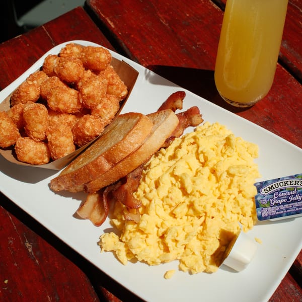 A breakfast plate with scrambled eggs, bacon, toast, tater tots, grape jelly, and orange juice.