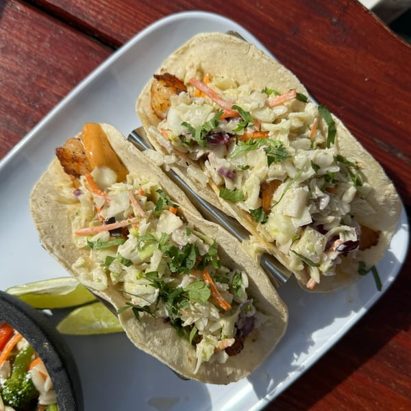 Two Cowgirl Kitchen fish tacos with slaw and cilantro on a white plate with lime wedges.