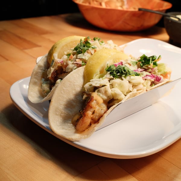 Two shrimp tacos with coleslaw, cilantro, and lemon wedges on a white plate.