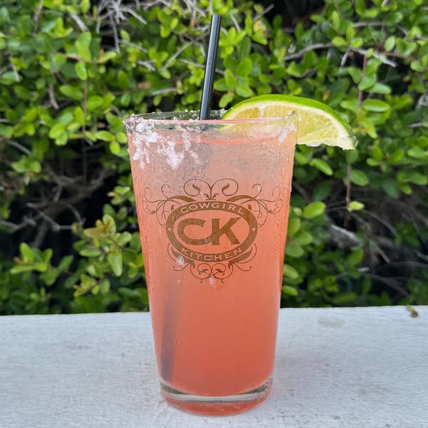 A Cowgirl Kitchen branded glass with a pink drink, salted rim, lime, and straw against greenery.