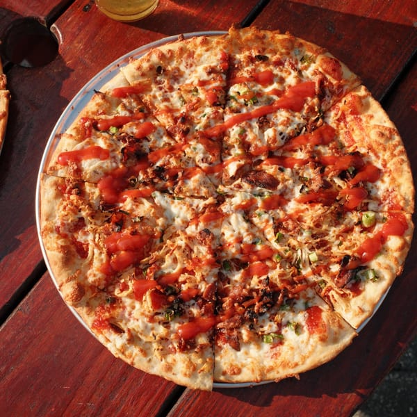 A Cowgirl Kitchen pizza with cheese, bacon, green onions, and red sauce on a wooden table.